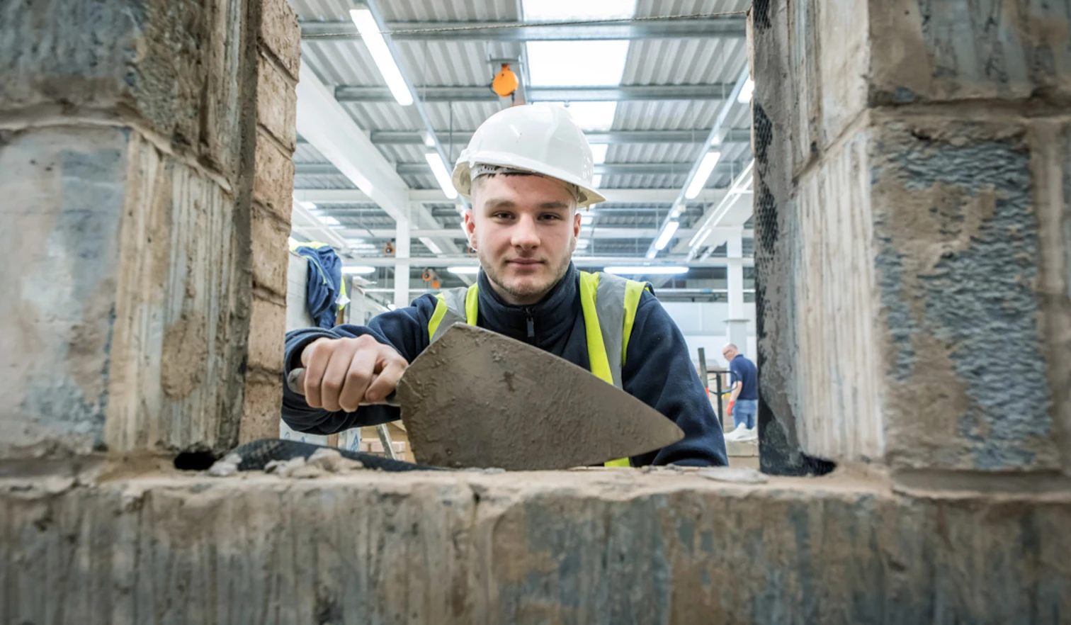 A brickwork student wearing a safety helmet and high-visibility vest using a trowel to apply mortar, viewed through an opening in a brick structure. A brickwork student wearing a safety helmet and high-visibility vest using a trowel to apply mortar, viewed through an opening in a brick structure.